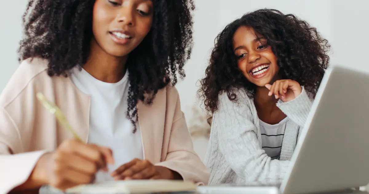 A mother and daughter with curly hair share a joyful moment as the mother writes in a planner, while the daughter smiles and looks on, highlighting the importance of staying organized for busy moms.