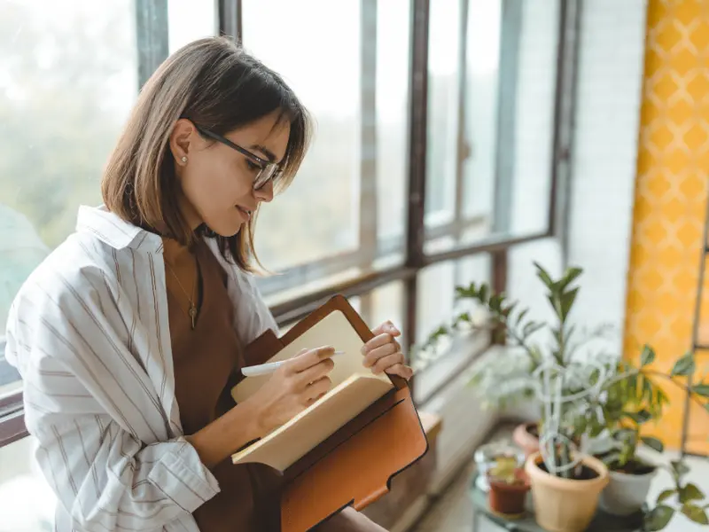 Woman next to window, writing in a notebook.