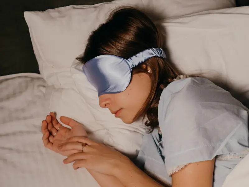 Woman sleeping in bed on her side wearing light blue silk eye mask. 