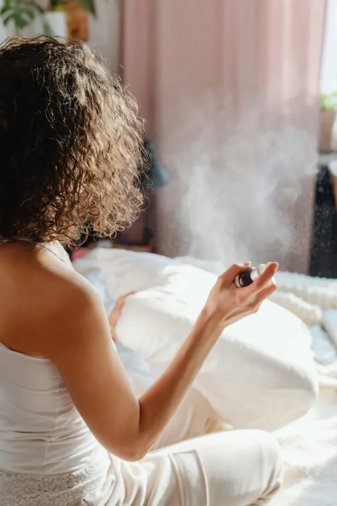 A woman sitting on a bed in a softly lit room, spraying a sleep relaxation spray onto her pillows.