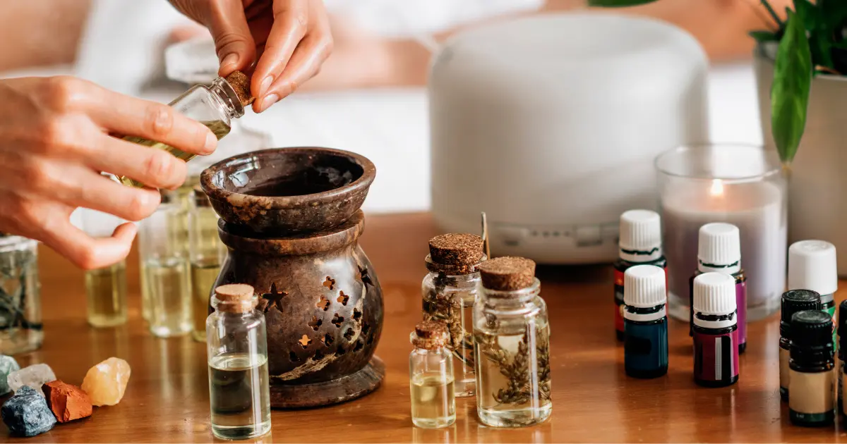 A person is carefully pouring essential oil into a rustic stone diffuser surrounded by various small glass bottles of oils, a lit candle, and crystals on a wooden surface. In the background, there is an ultrasonic diffuser, contributing to a serene, spa-like atmosphere.