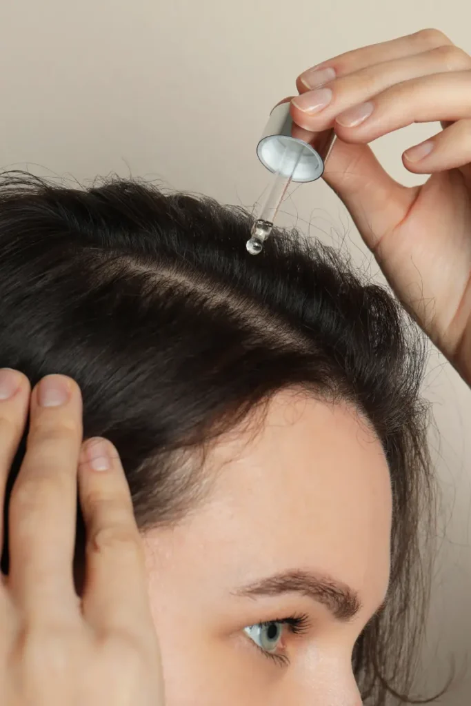 A close-up of a woman applying hair growth oil to her scalp using a dropper. 