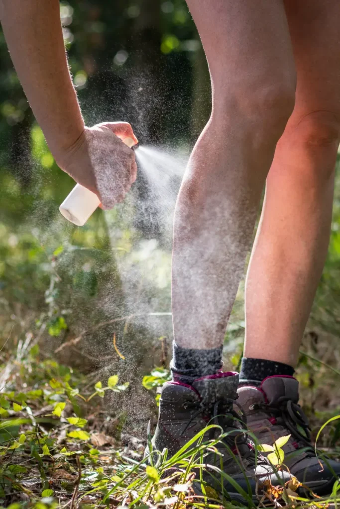 A person spraying insect repellent on their legs while standing outdoors, wearing hiking boots and socks. The spray is visible as a fine mist covering the skin.