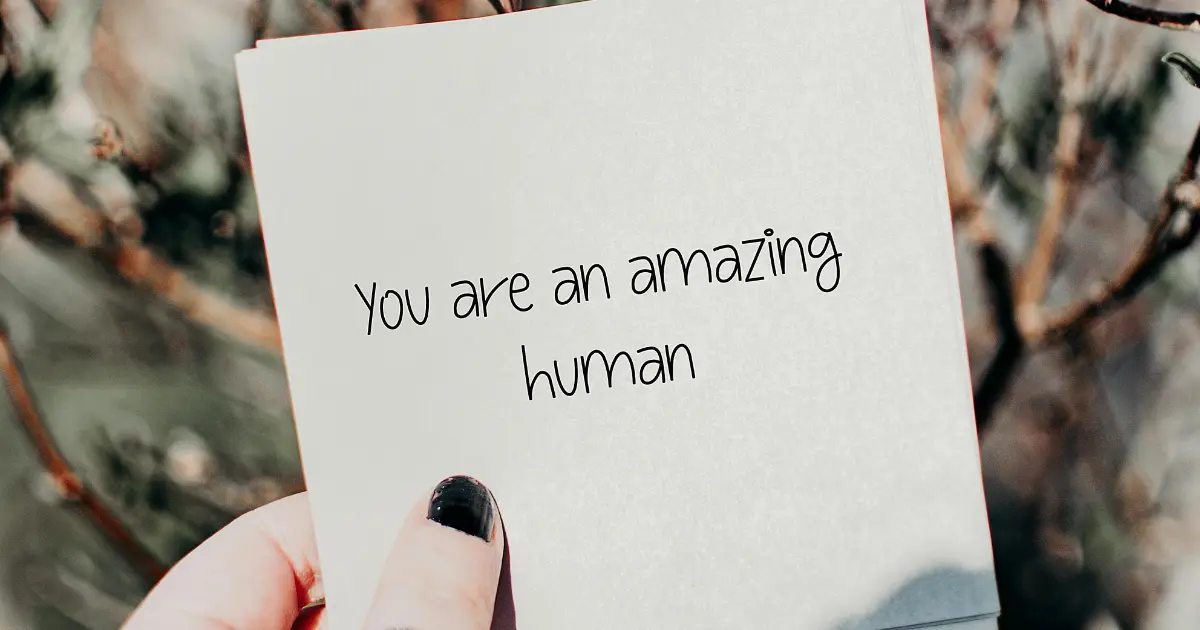 The image shows a close-up of a person's hand holding a small card with the text "You are an amazing human" written on it. The background is slightly blurred, showing outdoor foliage, giving the image a warm, uplifting feel. The person holding the card has black nail polish on their thumb.