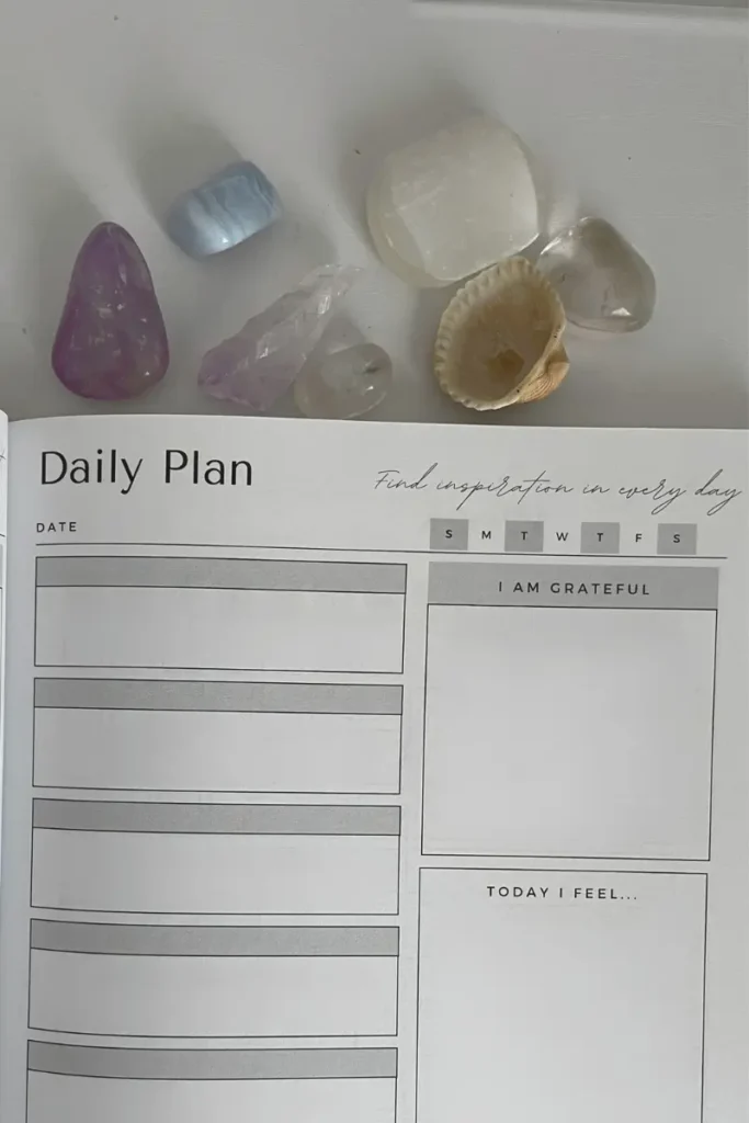 A daily planner open to a page with sections for the date, gratitude, feelings, and tasks. The page is accompanied by crystals and a seashell, with a motivational quote at the top that reads, "Find inspiration in every day," adding a touch of mindfulness to the planning process.