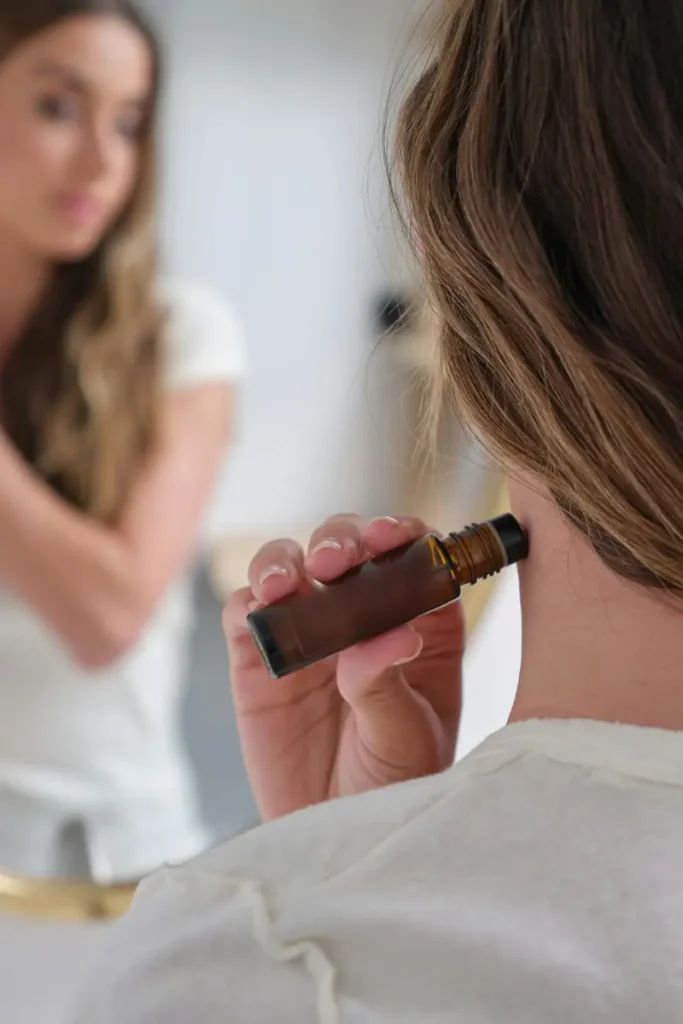 A woman applying a pain relief roller to the back of her neck while looking at herself in a mirror, with a focus on the hand holding the roller and the neck area.