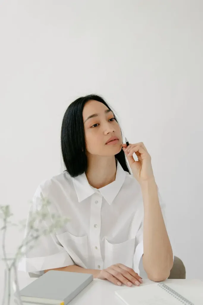 A woman in a white shirt sits thoughtfully at a desk, holding a pen near her face as she contemplates her writing. There is a closed notebook and an open journal in front of her, a moment of self-reflection and planning.