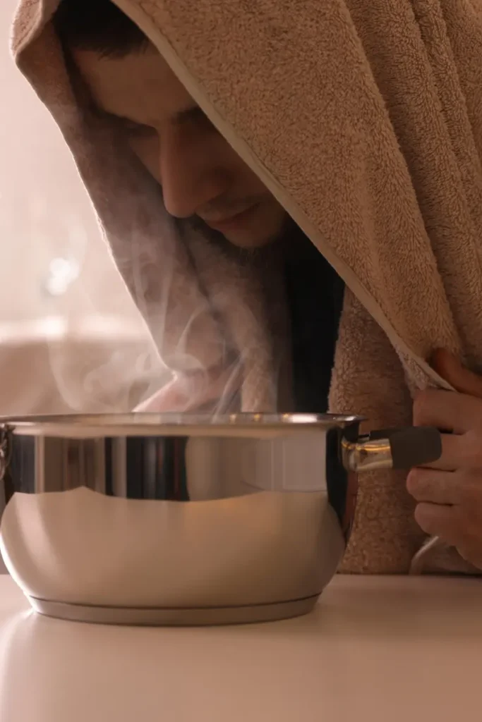 A person with a towel draped over their head, leaning over a steaming pot, inhaling the steam, for respiratory relief.