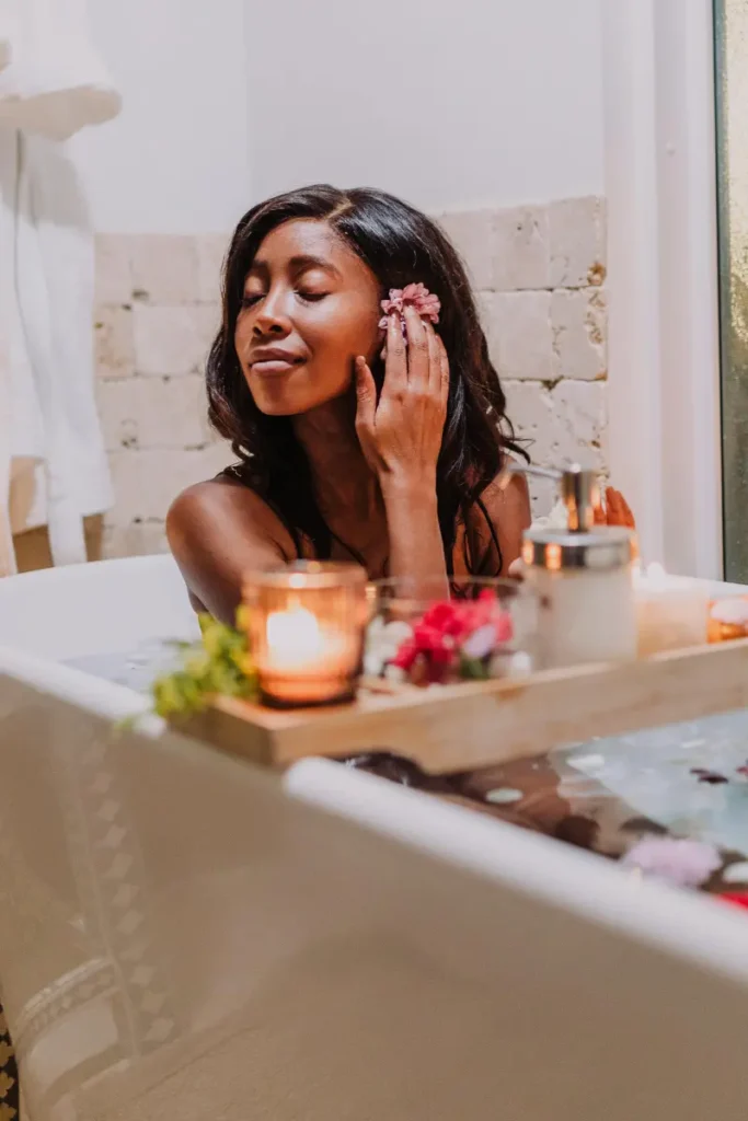 A woman relaxing in a bath surrounded by candles and flowers, gently touching a flower to her ear, creating a soothing and peaceful atmosphere.