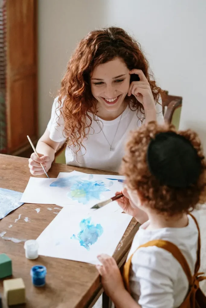 A smiling woman and child painting together at a table, using shades of blue on white paper.