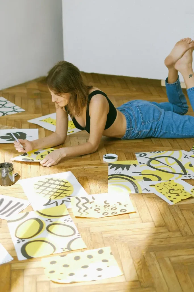 A woman lying on the floor, drawing abstract patterns in yellow and black on multiple sheets of paper around her.