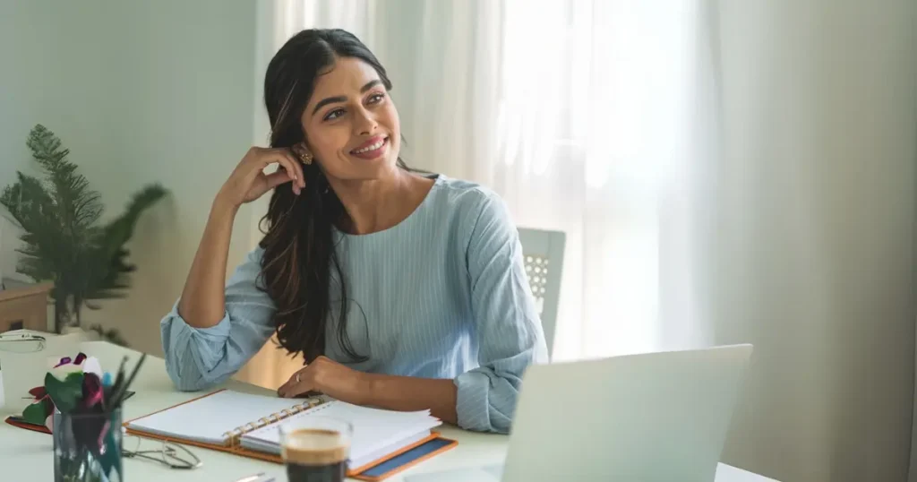 A mom entrepreneur in a light blue blouse sits at her desk, smiling thoughtfully while working on time management strategies. An open planner, a cup of coffee, and a laptop suggest a mindset shift toward overcoming time scarcity.