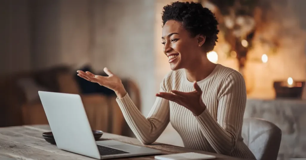 A smiling woman gestures while video chatting on her laptop, embodying conscious communication and confidence in a warm, inviting setting.