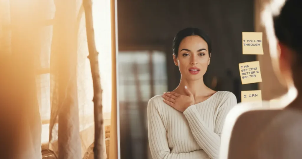 A woman stands in front of a mirror with her hand on her heart, speaking affirmations written on sticky notes: "I follow through," "I am getting better & better," and "I can." The daily affirmations rewire your brain.